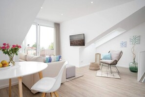 Une pièce moderne et bien éclairée, avec un canapé blanc, une table à manger agrémentée d’un vase de fleurs, une chaise et une vue sur l’extérieur grâce à une grande fenêtre.