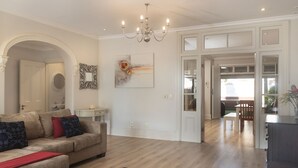 Een ruime woonkamer met een beige bank, een salontafel en een kroonluchter. Door een open deur is een eethoek met een tafel en stoelen zichtbaar.