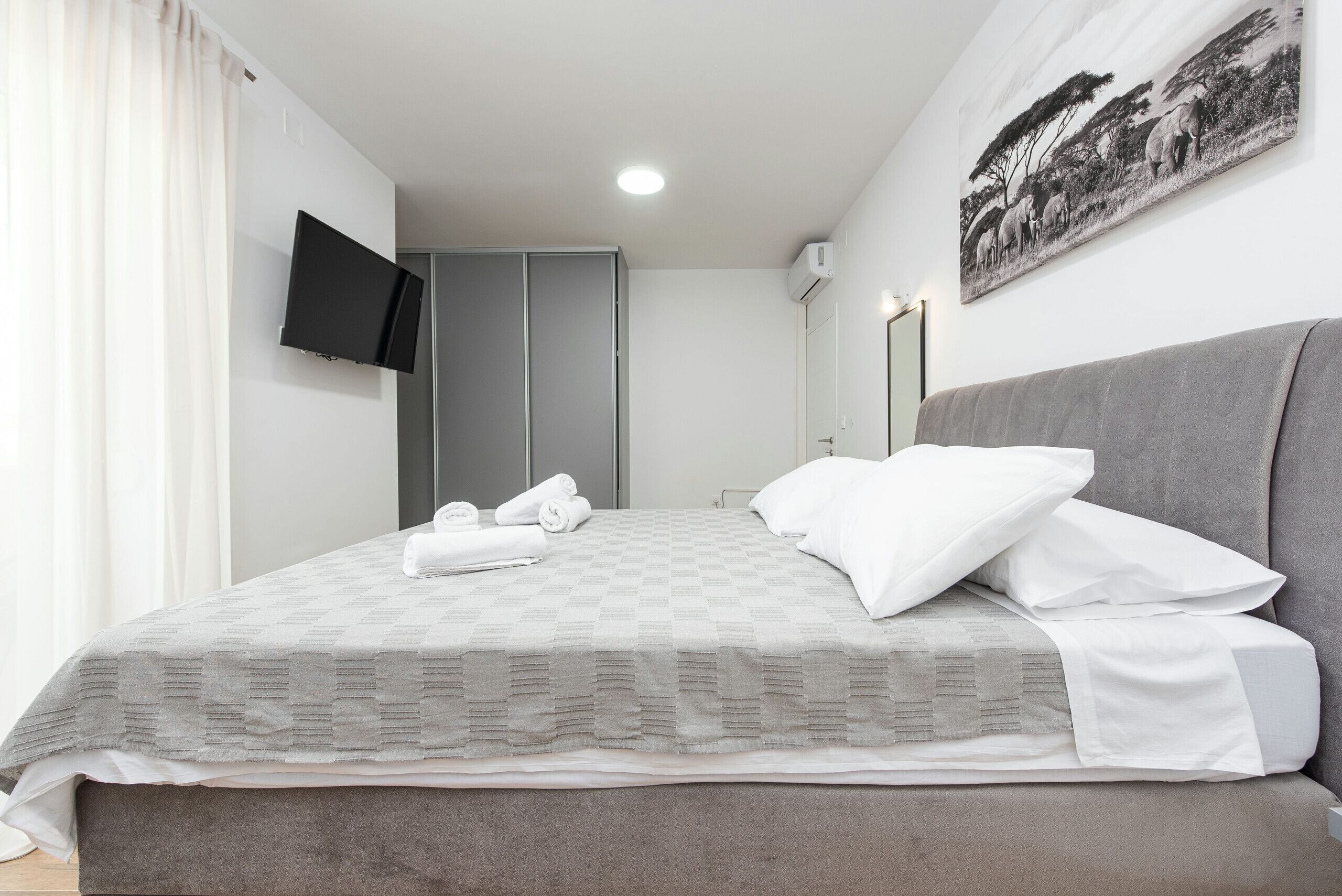 A neatly made bed with white linens, a grey headboard, and a mounted flat-screen TV on the wall.