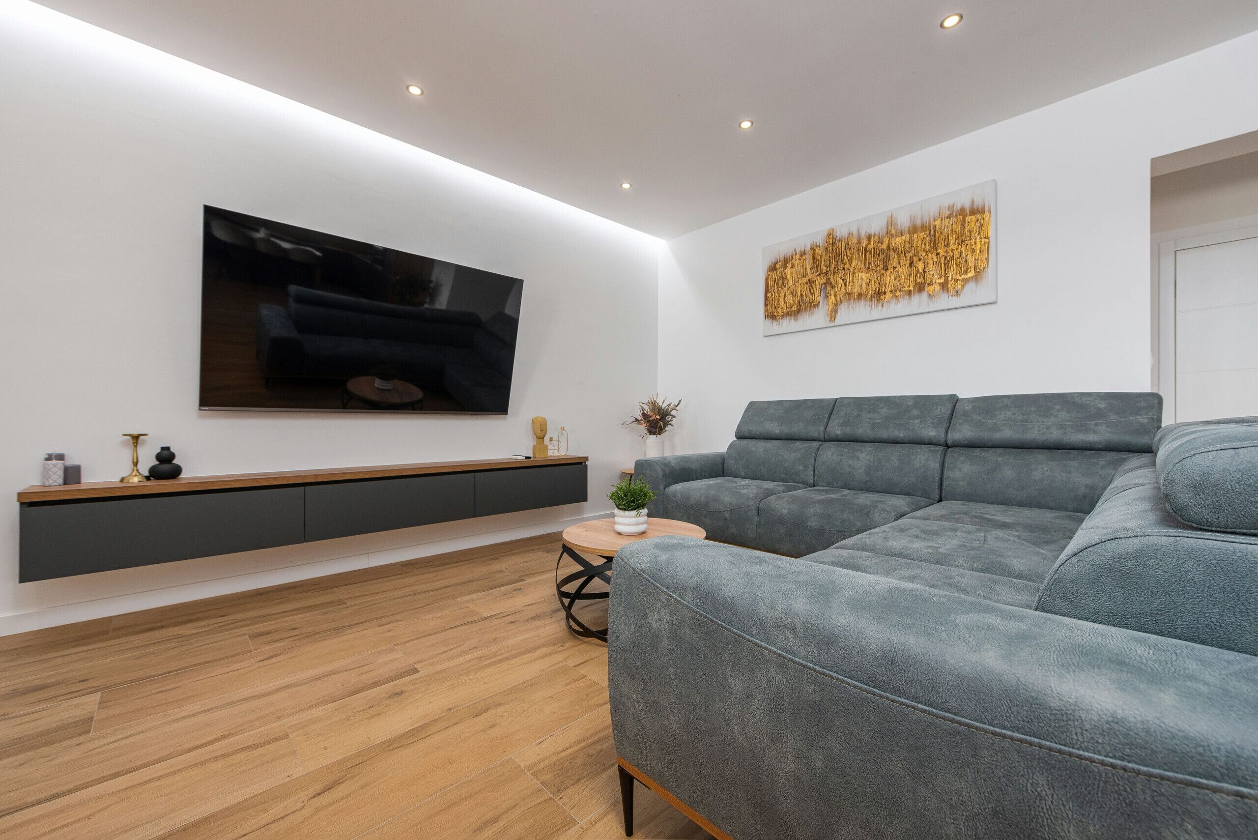 A modern living room with a grey sofa, a flat-screen TV mounted on the wall, a wooden floor, and a minimalist console table.