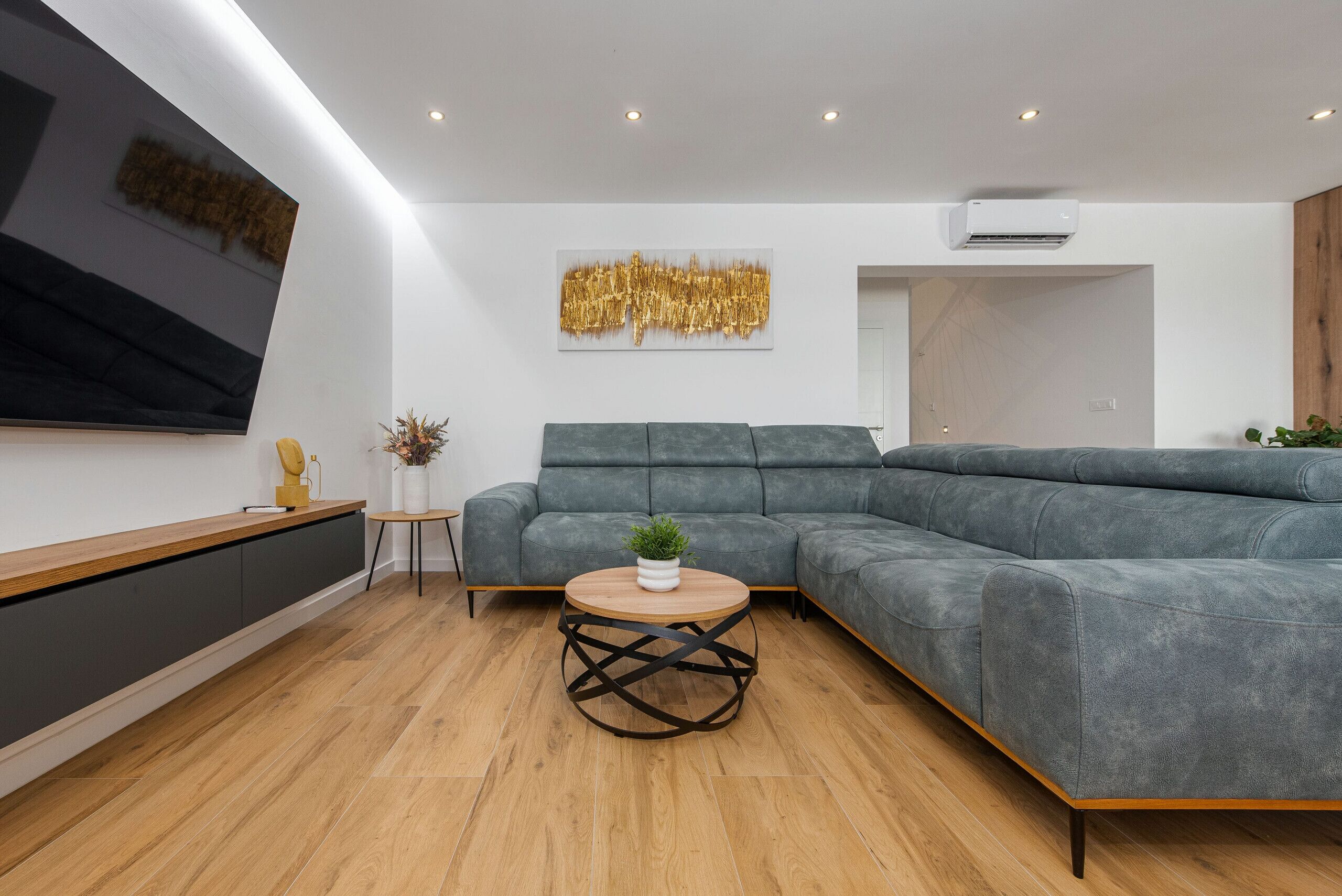 A modern living room with a grey sectional sofa, a round coffee table with a plant, a flat-screen TV mounted on the wall, and an air conditioning unit.