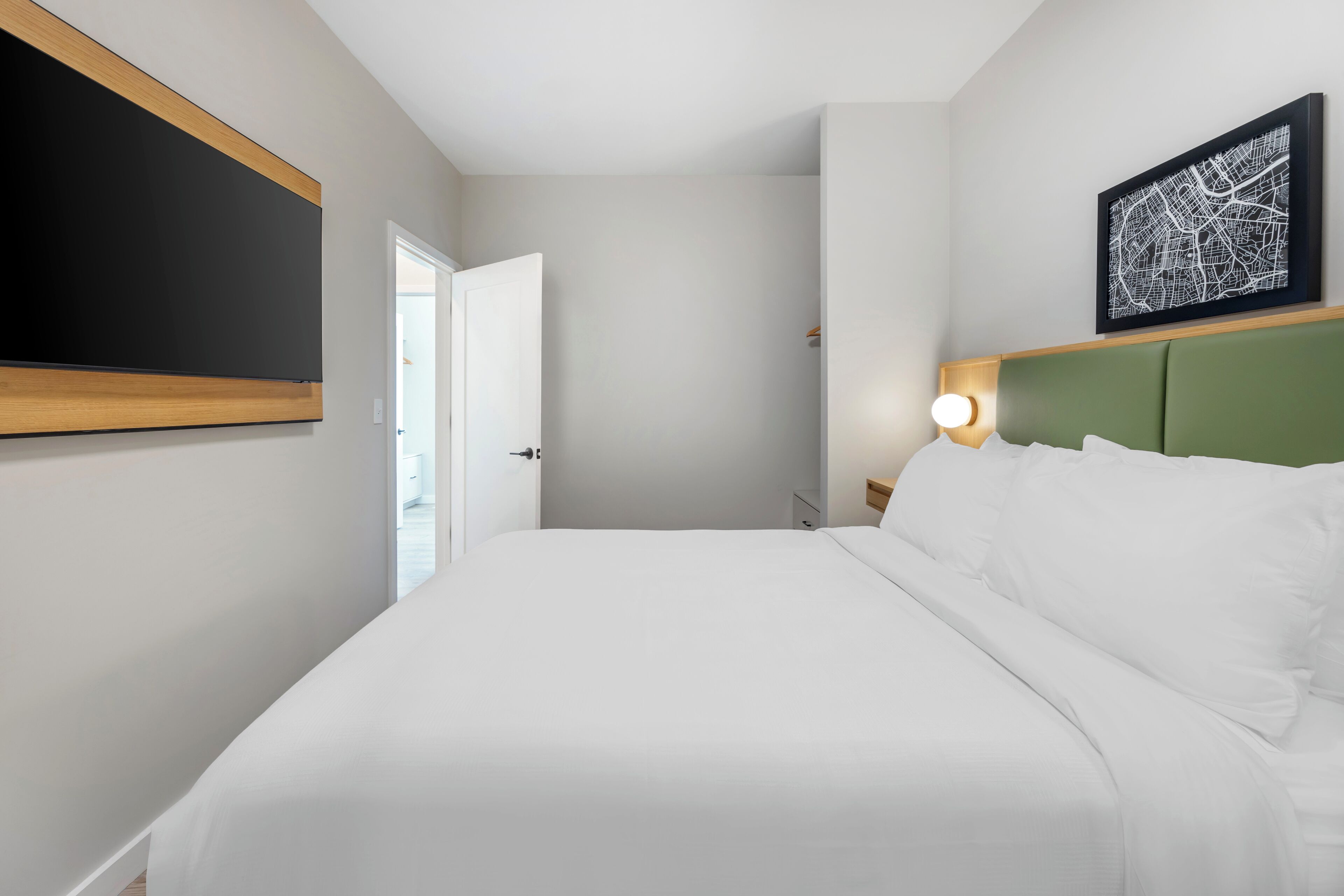 A hotel room with a large bed, a green headboard, a wall-mounted flat-screen TV, and a framed map on the wall.