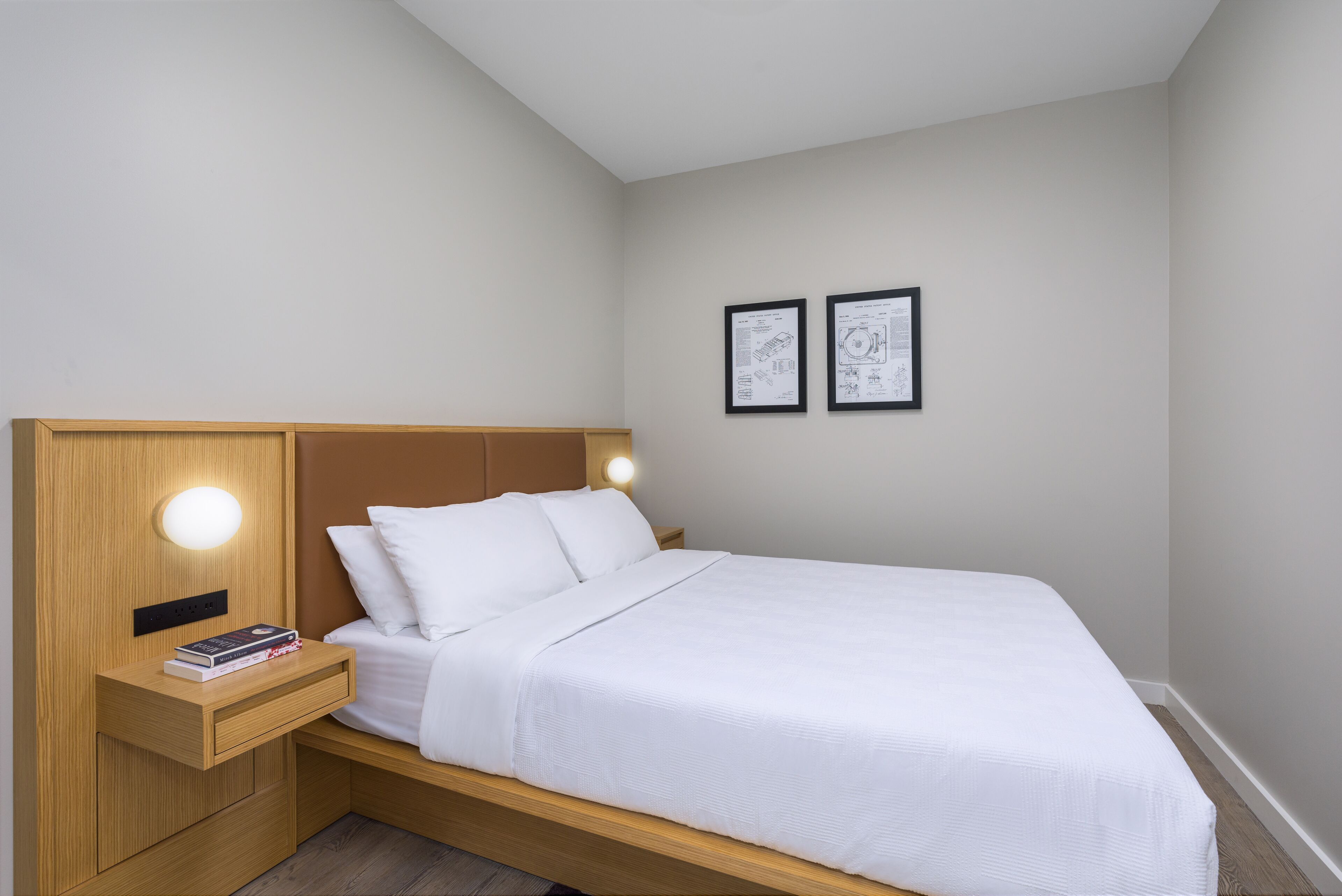 Una cama bien hecha con cabecera y mesita de noche de madera, una pequeña lámpara de noche y dos cuadros enmarcados en la pared.