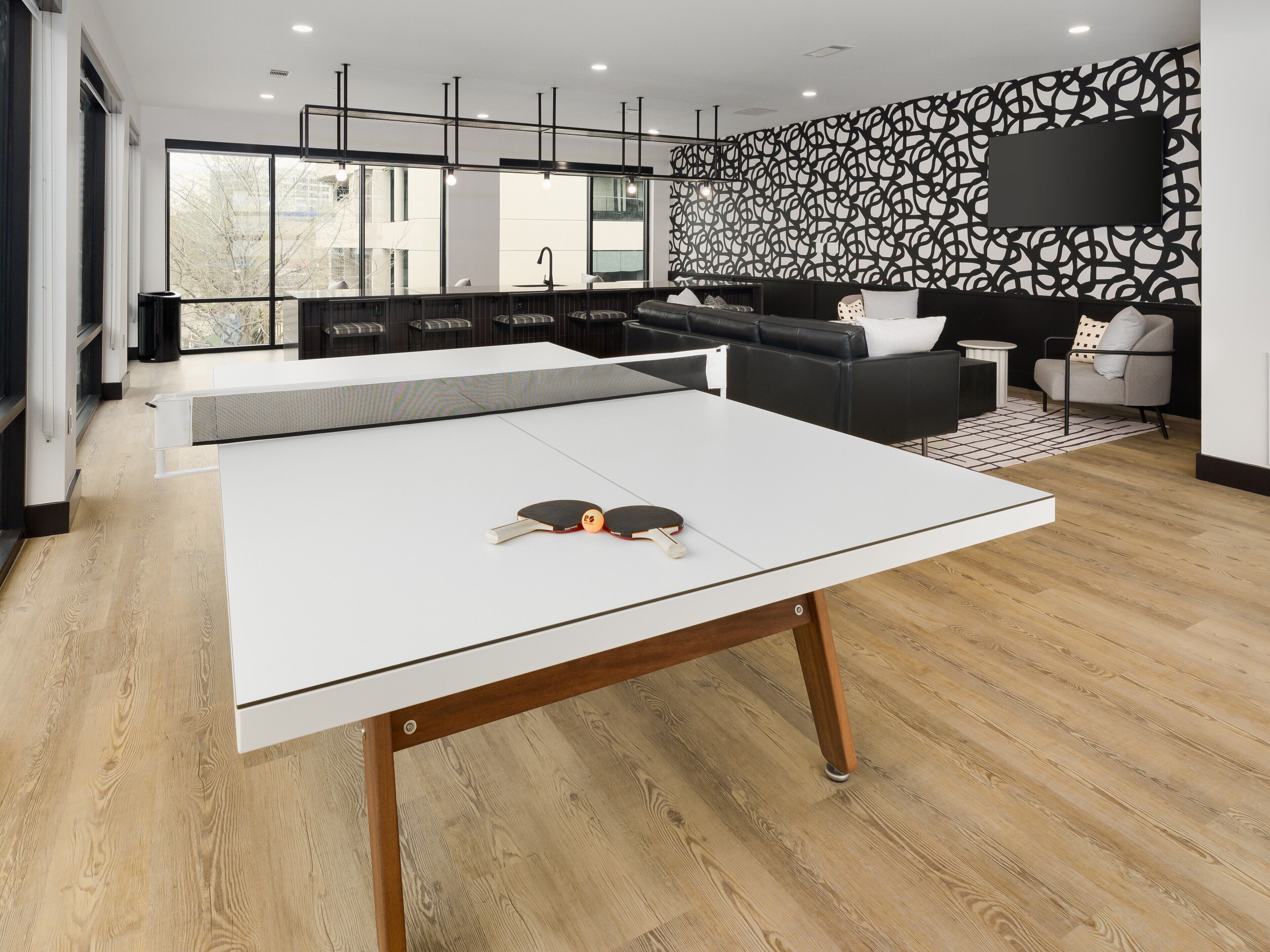 A modern living room with a ping pong table, a sofa, and a TV mounted on a wall with a geometric pattern.