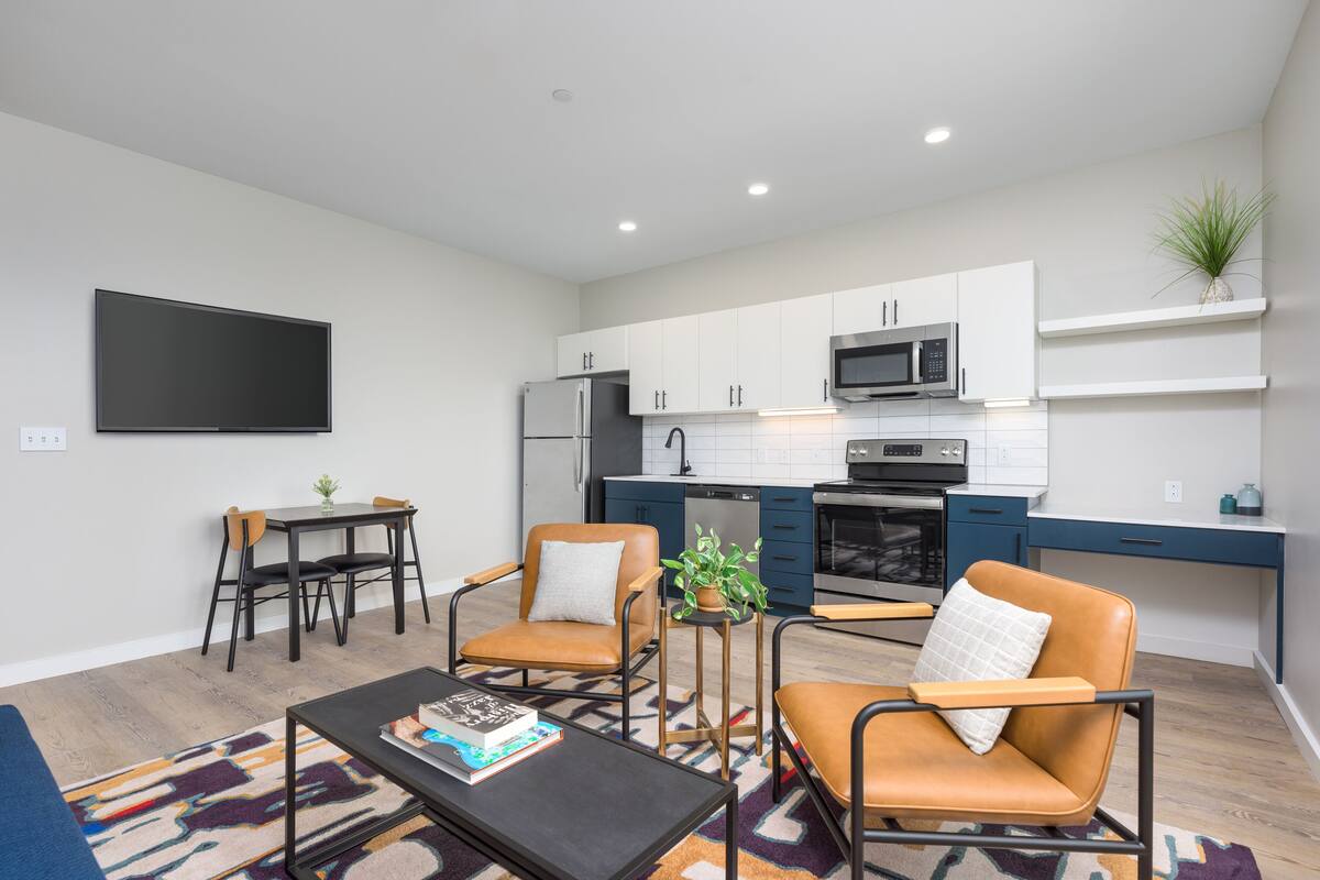 A modern kitchen with white cabinets, a microwave, and a dishwasher. There is a dining area with a table and chairs, and a living room with a sofa and a coffee table.