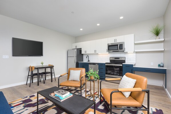 A modern kitchen with white cabinets, a microwave, and a dishwasher. There is a dining area with a table and chairs, and a living room with a sofa and a coffee table.