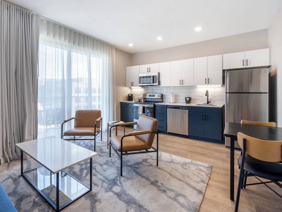 A modern kitchen with stainless steel appliances, a dining area with a glass table and chairs, and a large window with sheer curtains.