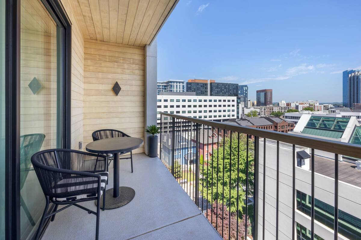 A balcony with a view of a cityscape, featuring a table and chairs, a potted plant, and a railing.