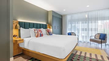 Ein modernes Hotelzimmer mit einem großen Bett, einem Nachttisch aus Holz, einer Schreibtischlampe, einem Sessel und einem großen Fenster mit transparenten Vorhängen.