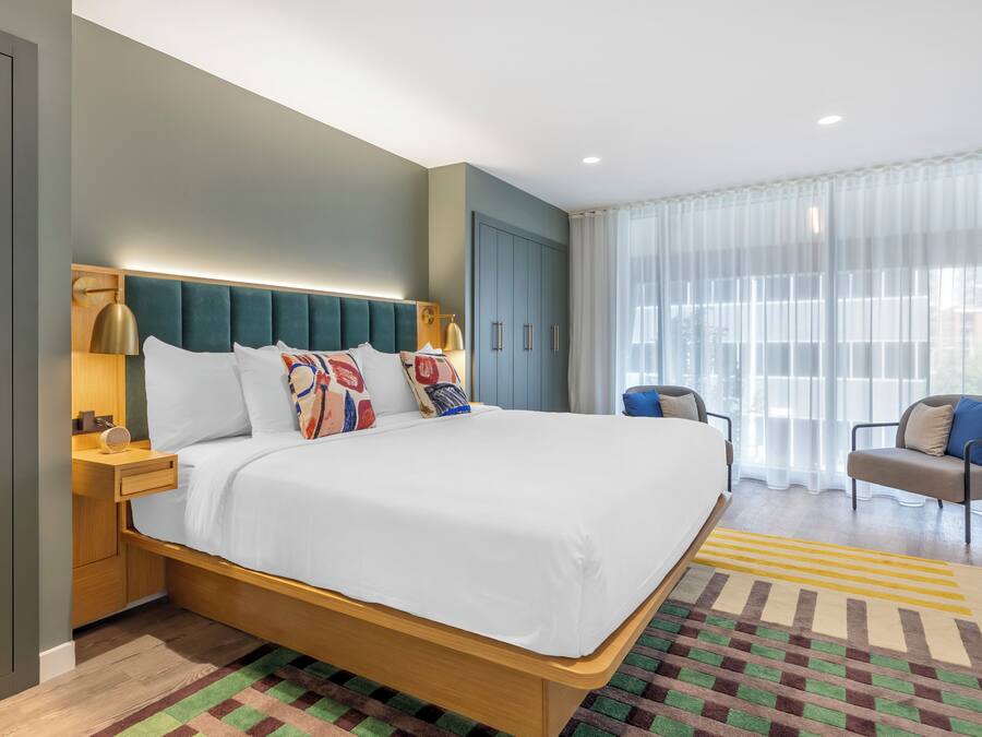 Ein modernes Hotelzimmer mit einem großen Bett, einem Nachttisch aus Holz, einer Schreibtischlampe, einem Sessel und einem großen Fenster mit transparenten Vorhängen.