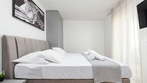 Ein modernes Schlafzimmer mit einem großen Bett, einem Nachttisch, einem an der Wand befestigten Fernseher und einem Fenster mit durchsichtigen Vorhängen.