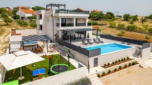 Ein modernes Haus mit zwei Etagen, einem Swimmingpool, Sitzgelegenheiten im Freien und einem Trampolin.