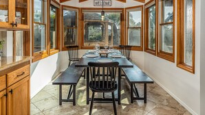 Un coin repas avec une table en bois dressée pour quatre personnes, entouré de grandes fenêtres et d’une pancarte indiquant « BIENVENUE ».