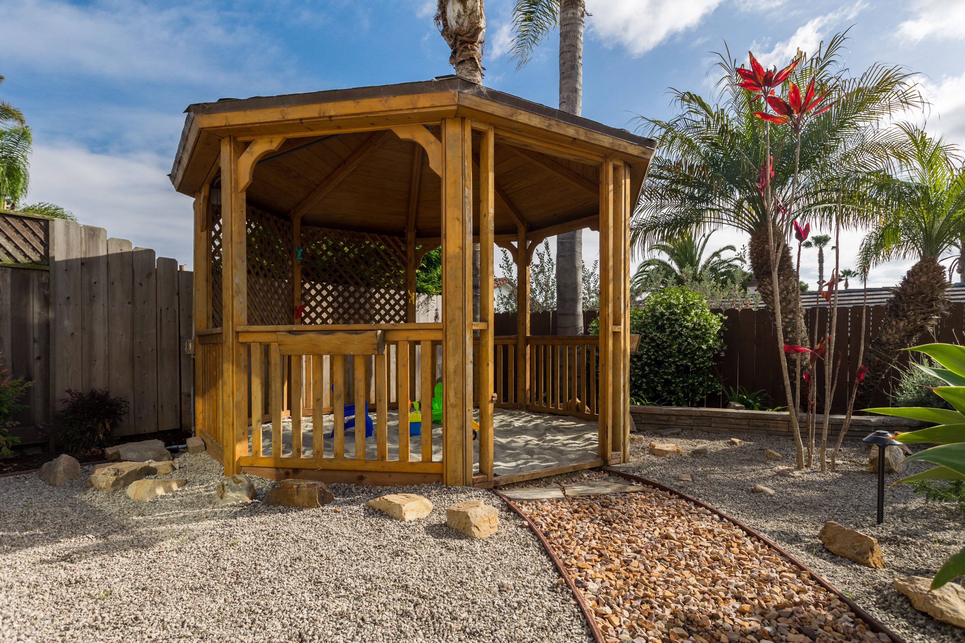A wooden gazebo with a lattice roof, surrounded by gravel and rocks, and palm trees in the background.