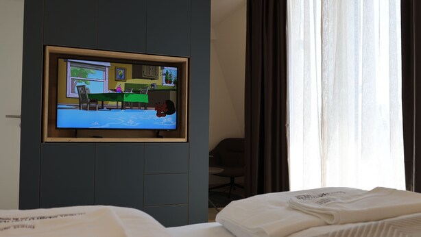 Ein Hotelzimmer mit Bett, Schreibtisch mit Stuhl, Wandfernseher und einem Fenster mit Vorhängen.