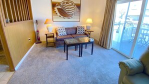 Uma sala de estar com sofá, duas mesas laterais com luminárias, uma mesa de centro e uma janela grande com cortinas.