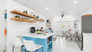 Une cuisine moderne avec un îlot central bleu, des tabourets de bar blancs, des étagères en bois et un coin repas avec des chaises en osier.