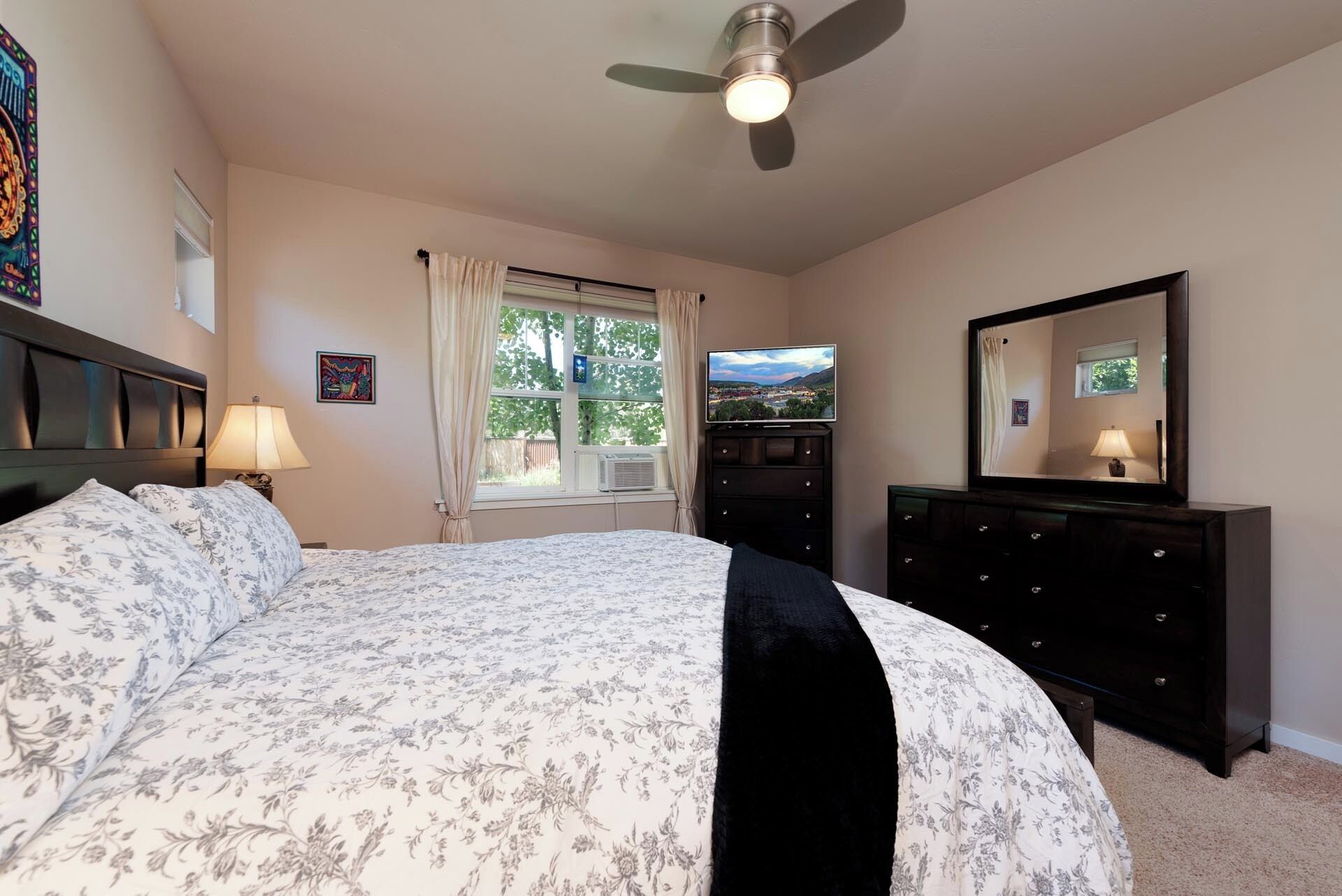 A bedroom with a bed, a dresser, a ceiling fan, a window with curtains, and a TV on a dresser.