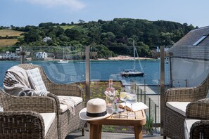 A wicker lounge chair with a cushion, a small table with a hat, a bottle, and glasses, overlooking a marina with boats and a hilly landscape. - The Lofthouse -  a luxury waterside apartment (Salcombe)
