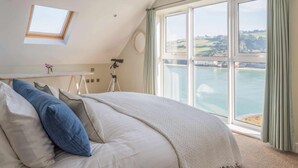 Een slaapkamer met een groot raam dat uitkijkt op een waterpartij en een brug, een telescoop en een klein tafeltje met een bloem.