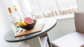 A table with a bowl of citrus fruits and a bottle of liquor, a wooden cutting board, and a window with the text "The Loft House". - The Lofthouse -  a luxury waterside apartment (Salcombe)