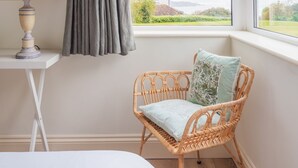 Un dormitorio con cama, una silla de mimbre con cojín, una mesita de noche con lámpara, una ventana que ofrece vistas a una casa y vegetación, y una cortina a rayas.