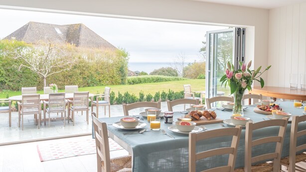 Comedor abierto con vistas al mar y a la montaña, con una mesa dispuesta para el desayuno y un florero con flores.
