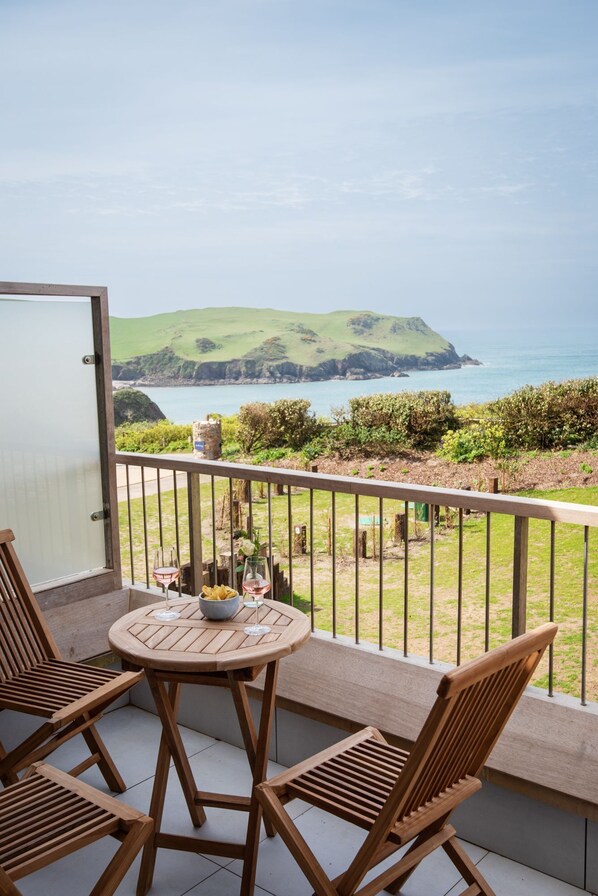 A balcony with a wooden table and chairs, overlooking a coastal landscape with hills and the sea.