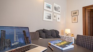 Een hotelkamer met een laptop op een tafel, een boek en een pen. Er hangen afbeeldingen in lijsten aan de muur en een lamp staat op een bijzettafeltje.