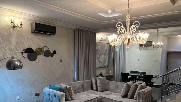 A modern living room with a sectional sofa, a round ottoman, a chandelier, and a dining area in the background. - Lovely nb 3-bed House in Opebi (Lagos)