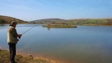 A man fishing by a calm lake with a distant island and rolling hills in the background.
