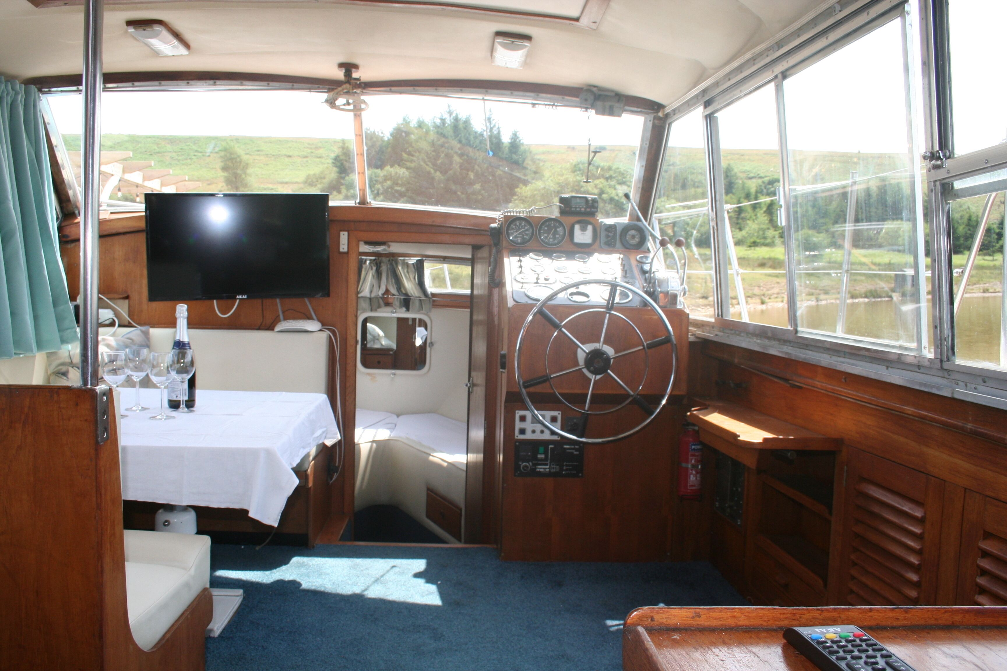 Interior de un barco con timón, panel de control y una mesa con vasos y una botella.