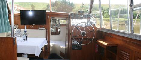 Interior de un barco con timón, panel de control y una mesa con vasos y una botella.