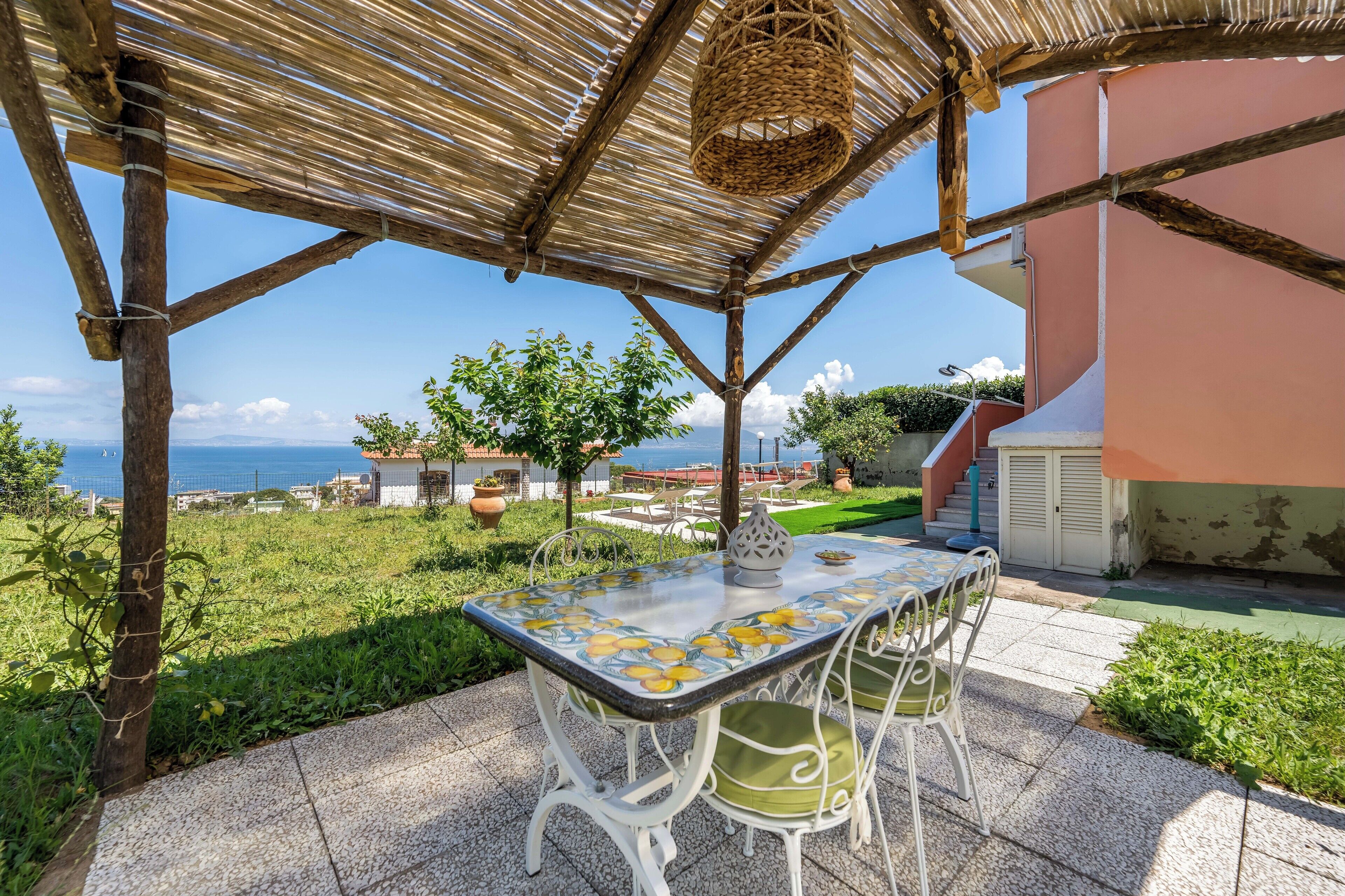 An outdoor patio with a table and chairs, a woven basket hanging from the ceiling, and a view of the sea and buildings in the background.