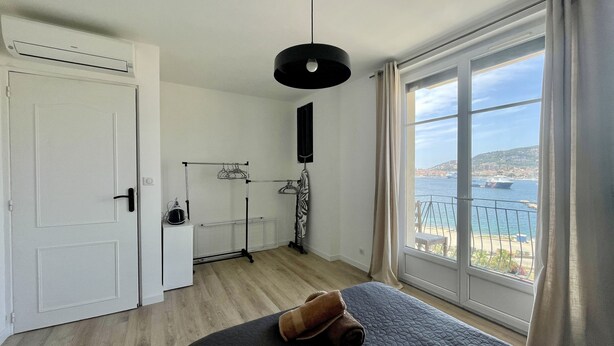 Ein Schlafzimmer mit Bett, Heizkörper, Pendelleuchte, Fenster mit Meerblick und einer Tür.