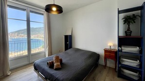 Ein Schlafzimmer mit einem Bett, einem Nachttisch mit Lampe, einem Ablagetisch mit Handtüchern und Meerblick durch ein großes Fenster.