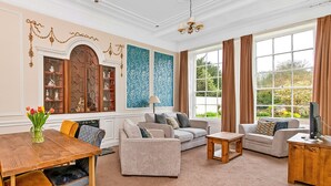 A spacious living room with a wooden coffee table, two sofas, a TV stand, and a large window with curtains. - Hambleton Hall Apt (Thirsk)