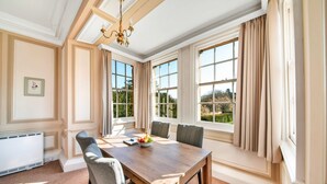A dining room with a wooden table, grey upholstered chairs, and a view of the outdoors through large windows.