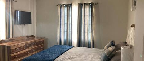 A bedroom with a bed, a wooden dresser, a television, and curtains.
