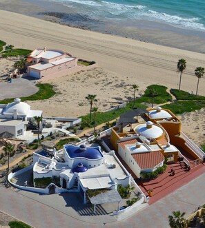 A coastal resort with multiple buildings, including a large white structure with blue domes, a yellow building, and a pink building. There is a beach and palm trees in the background.