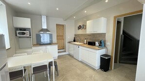 A compact kitchen with a microwave, sink, and coffee maker. There is a dining table with chairs and a wooden door leading to another room. - Maison de Village, 3 Chambres, Fibre (Dunes)
