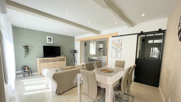 A modern living room with a dining area, a television, and a kitchen in the background. - Maison de Village, 3 Chambres, Fibre (Dunes)