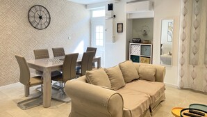 A modern living room with a beige sofa, a dining table with chairs, a clock on the wall, and a kitchen area in the background. - Maison de Village, 3 Chambres, Fibre (Dunes)