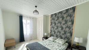 A bedroom with a bed, two bedside tables, a nightstand, a window with curtains, and a patterned wallpaper. - Maison de Village, 3 Chambres, Fibre (Dunes)