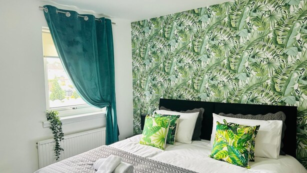 Een slaapkamer met een bed, twee kussens met tropische bladmotieven, een raam met turquoise gordijnen en een radiator.