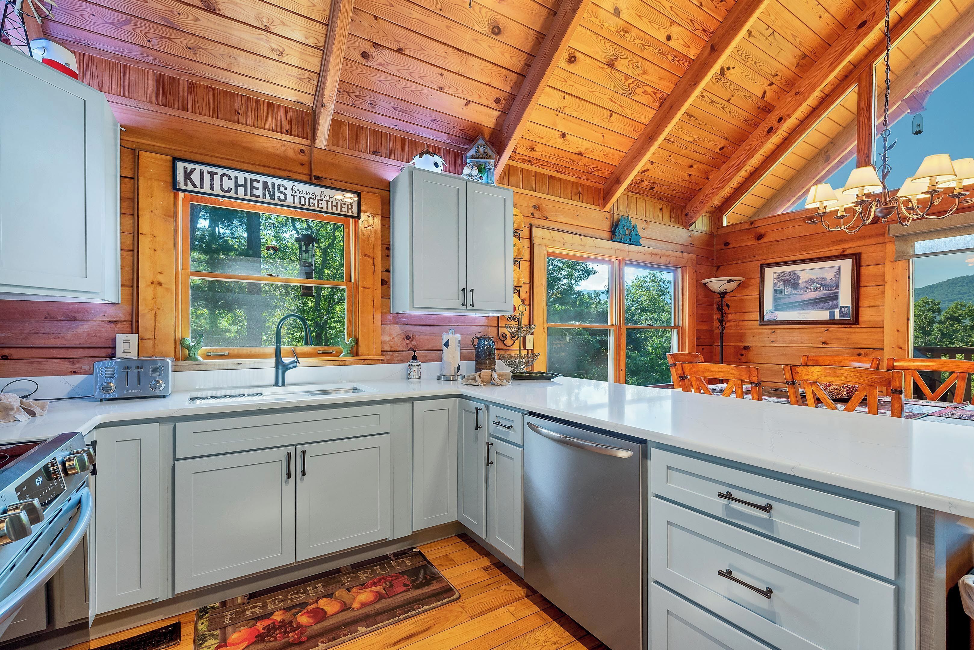 A kitchen with wooden beams, white cabinets, a dishwasher, and a window with a view of trees.