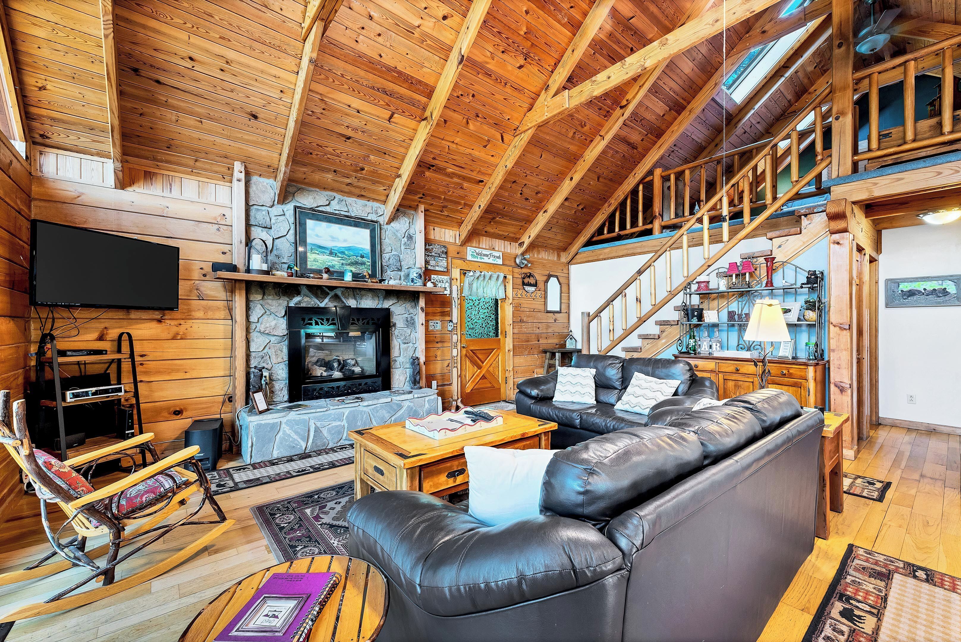 A log cabin interior with a stone fireplace, leather sofas, wooden floors, and a TV mounted on the wall.