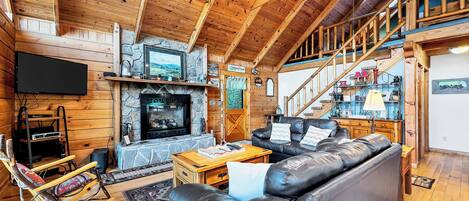 A log cabin interior with a stone fireplace, leather sofas, wooden floors, and a TV mounted on the wall.