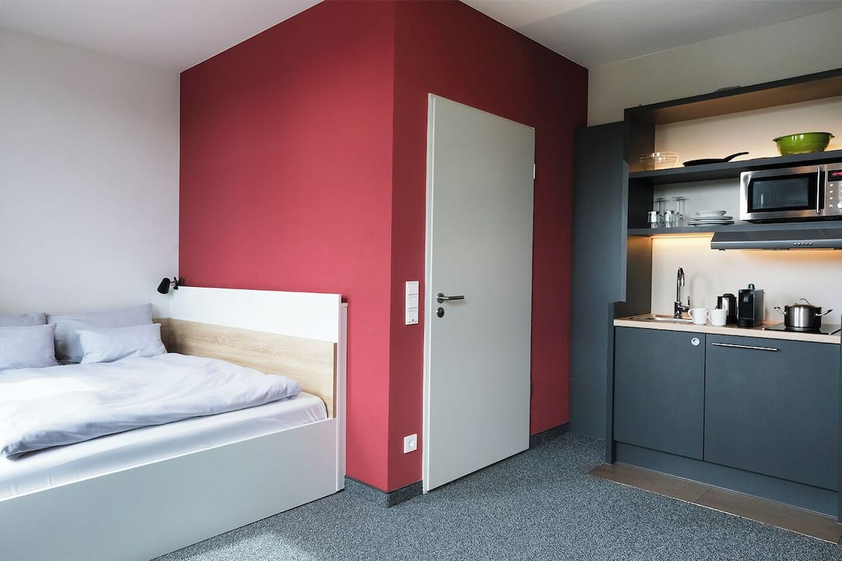 A compact room with a bed, a kitchenette, and a door.
