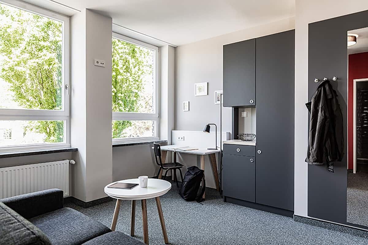 A compact room with a kitchenette, a small table with a chair, a radiator, and a window with a view of trees.